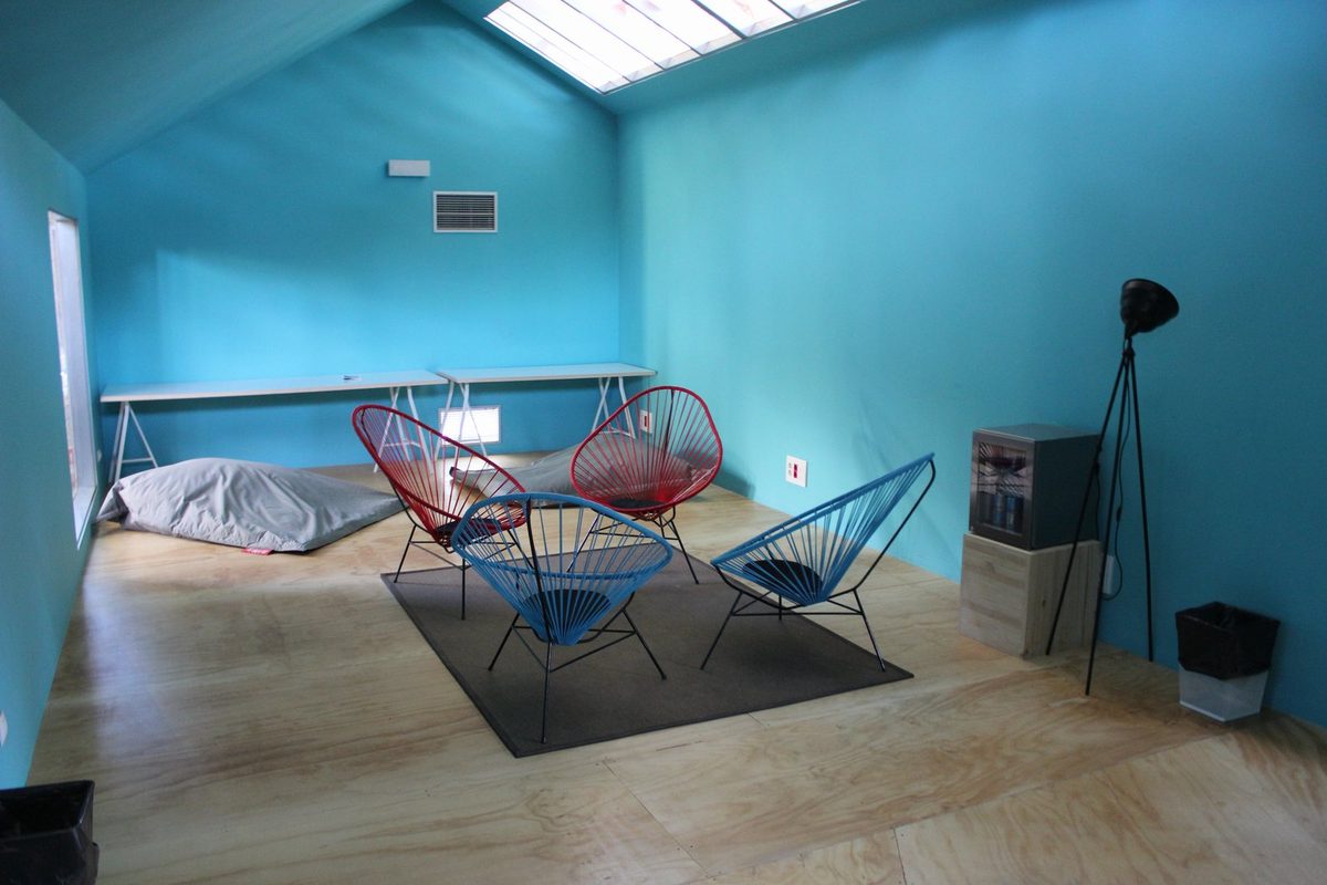 Séjour presque vide d'un module d'habitation aux murs turquoise, sol en bois et quatre fauteuils en fil métallique, tel que le voit un acheteur