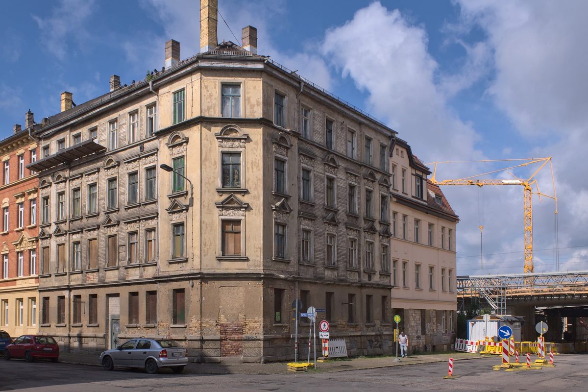 Unsaniertes Mehrfamilienhaus in Ecklage in Leipzig mit verwitterter Fassade und zugemauerten Fenstern, im Hintergrund ein Baukran an der Bahnbrücke