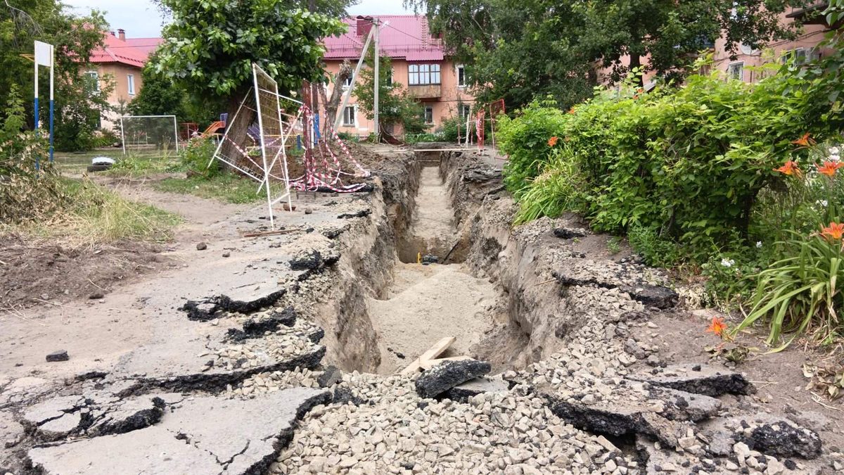 Langer Leitungsgraben quer durch einen asphaltierten Wohnhof, aufgebrochene Asphaltplatten und Absperrband, dahinter Wohnhäuser und ein Spielplatz