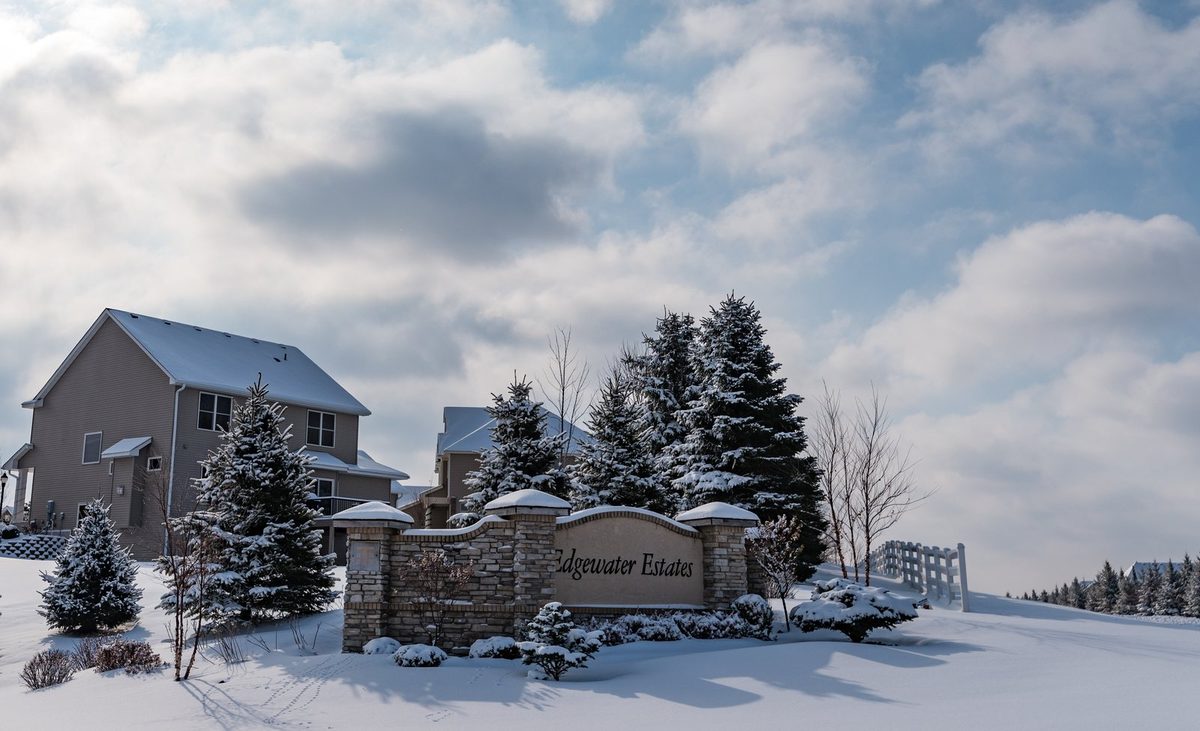 A for-sale sign in front of a suburban house with snow on the lawn and bare trees on a grey winter day