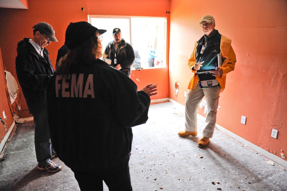 Home inspection in a gutted US room with stripped walls and a bare concrete floor, the inspector taking notes on a tablet while the owner looks on