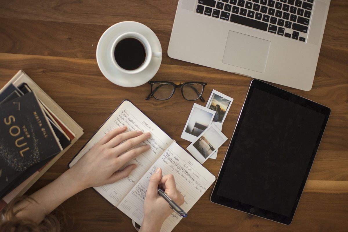 Vista dall'alto: una persona scrive a mano su un'agenda tra caffè, occhiali, laptop e tablet, scrivania tipica di chi prepara la vendita di casa